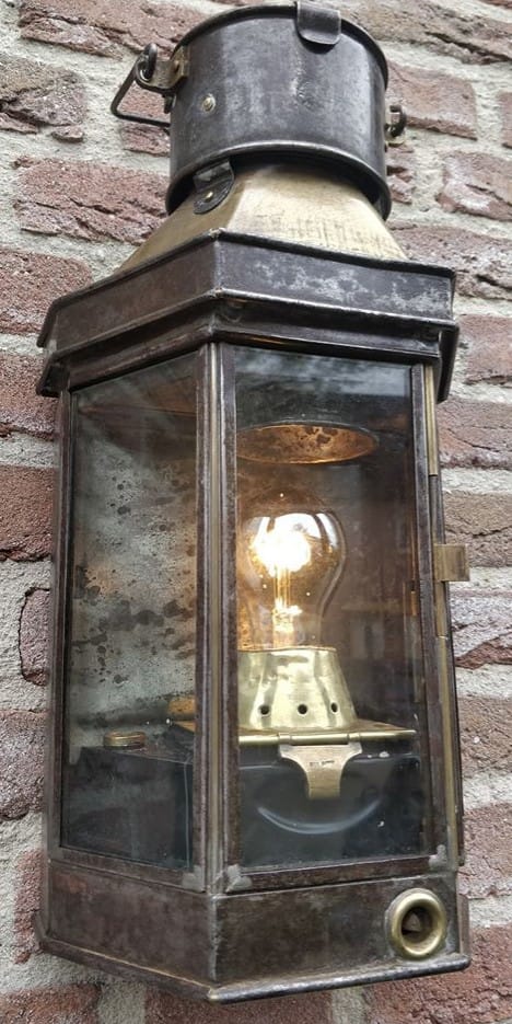 Scheepslamp van Alderson Gyde hangend aan een muur met brandende lamp